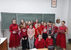 Grupa uczniów z klasy czwartej pozuje do zdjęcia w sali lekcyjnej, dwóch uczniów siedzi na podłodze, większa grupa dzieci stoi pod szkolną tablicą.